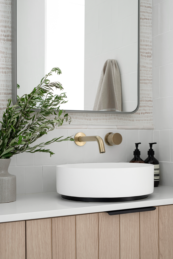 A bathroom vanity with a white sink and brass faucet. The Dashes wallpaper is on the walls. It's a small scale pattern in taupe that reads as a texture.