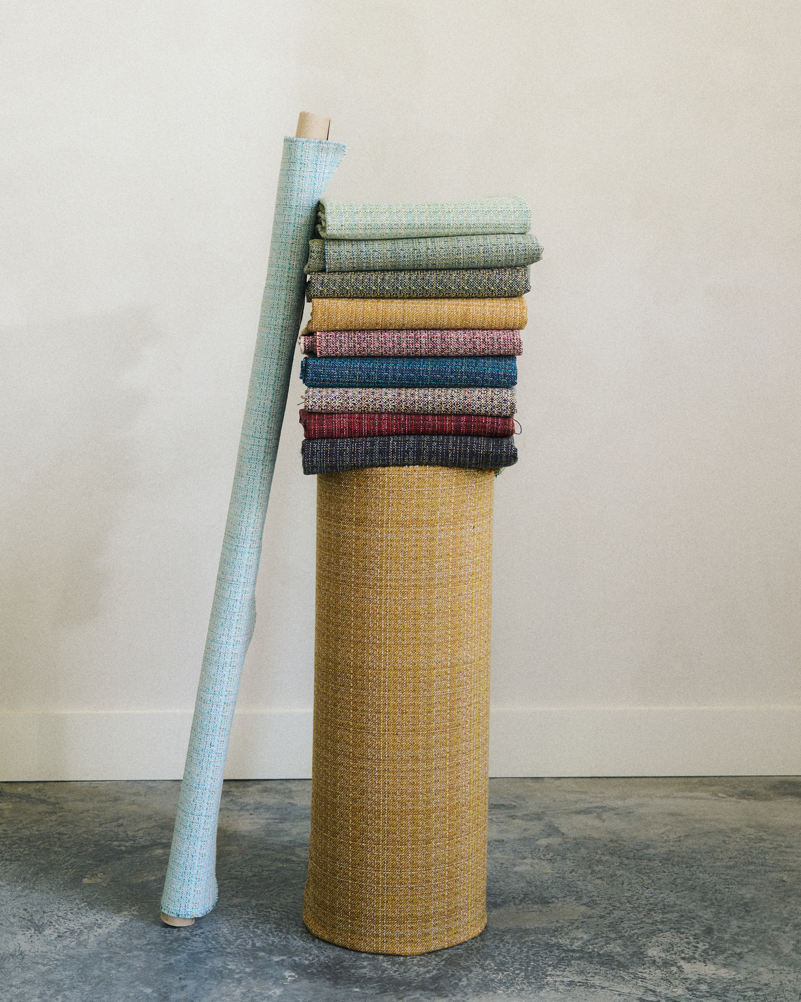 A pillar wrapped in a golden ochre fabric with a stack of fabrics on top of it in: blues, pink, purple, greens, yellows, and red. There is a roll of fabric in light blue leaning on the left. This is an upholstery cotton blend fabric.
